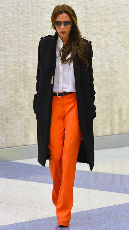 Βικτώρια Beckham wearing orange pants, white top, and black trench coat